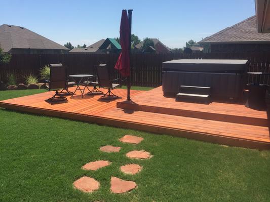 Backyard with a wooden deck featuring a covered hot tub, patio furniture, and a closed red umbrella, surrounded by lush green grass and a wooden fence.