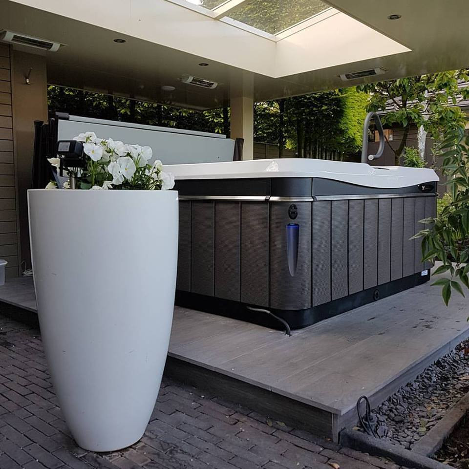 Stylish outdoor hot tub under a covered patio with a skylight, surrounded by greenery and modern décor, including a tall white planter with blooming flowers.