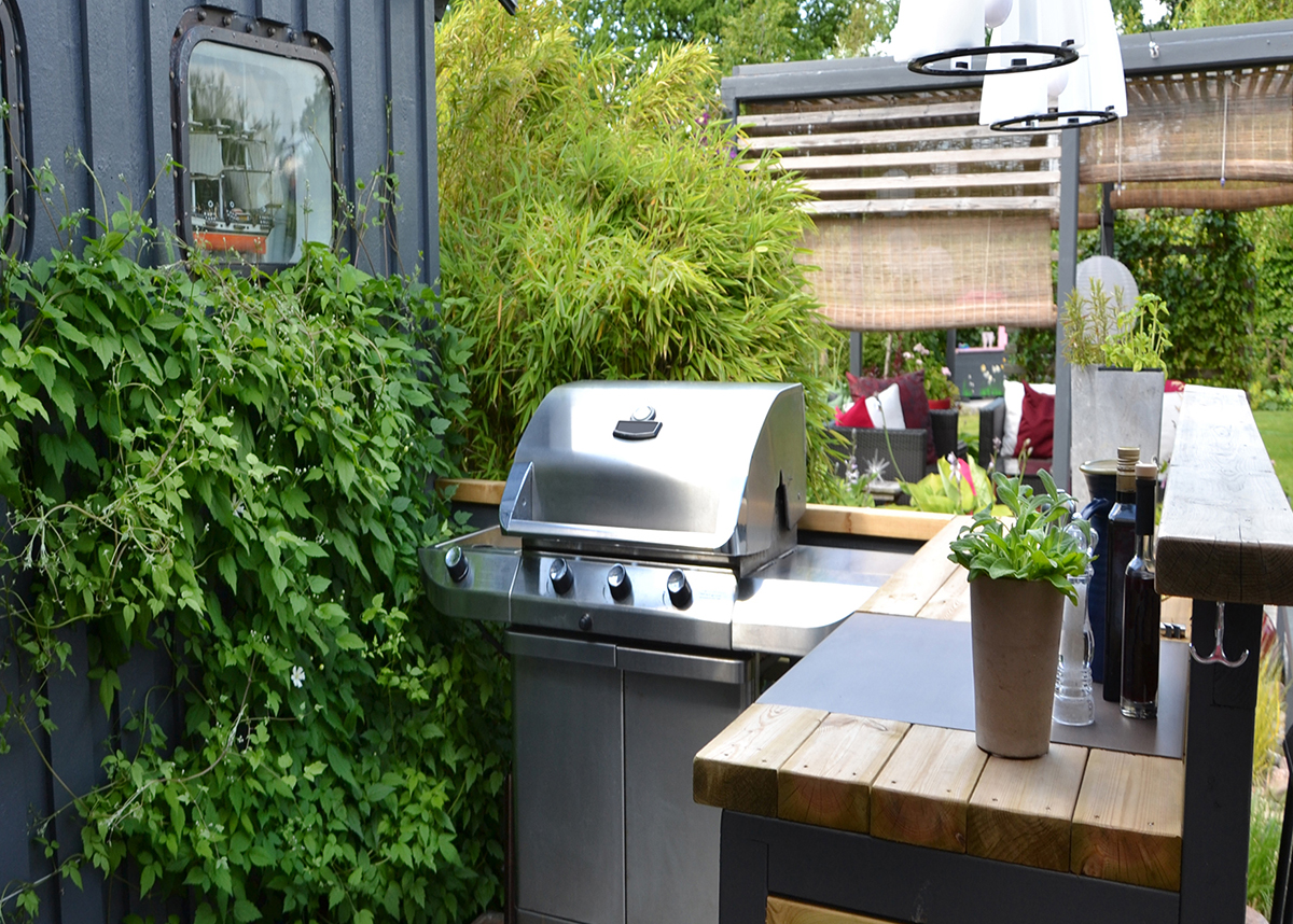 Outdoor lounging area with a grill. The area has comfortable seating, a grill, and a fire pit. The background is a lush green lawn.