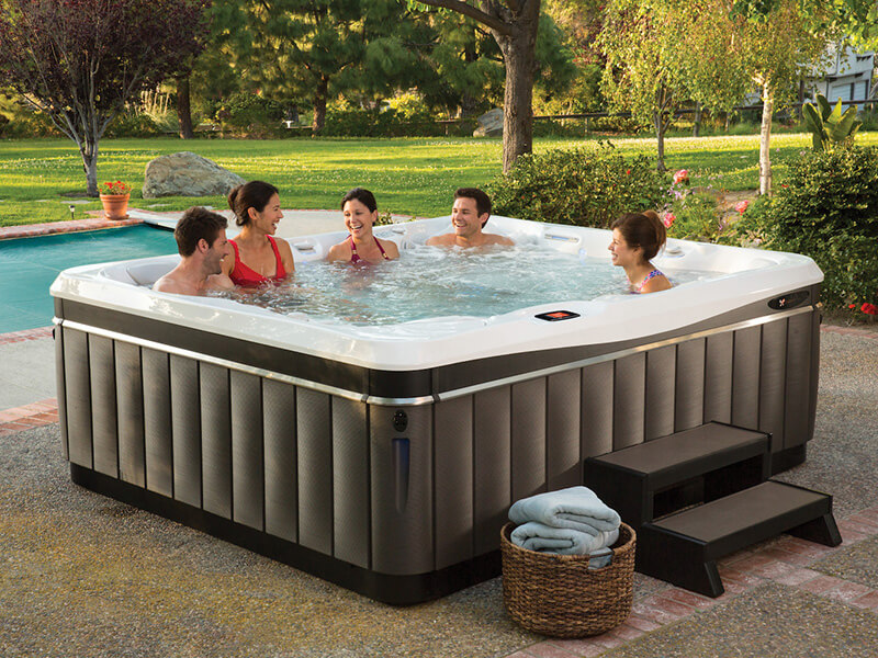 Group of five friends enjoying a conversation in a spacious outdoor hot tub, surrounded by lush greenery, a pool, and a well-manicured backyard.