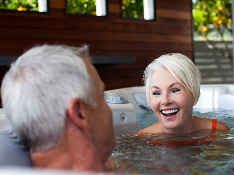 Smiling older couple enjoying a conversation while relaxing in a warm hot tub, with a modern wooden-paneled outdoor space in the background.