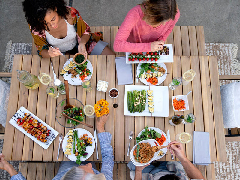 Overhead view of a group of people enjoying a vibrant outdoor meal at a wooden dining table, featuring grilled vegetables, fresh salads, and glasses of white wine.
