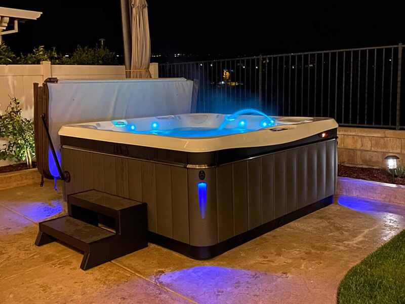 Modern outdoor hot tub illuminated with blue LED lights at night, featuring a water fountain and sleek design, set on a patio with ambient lighting.