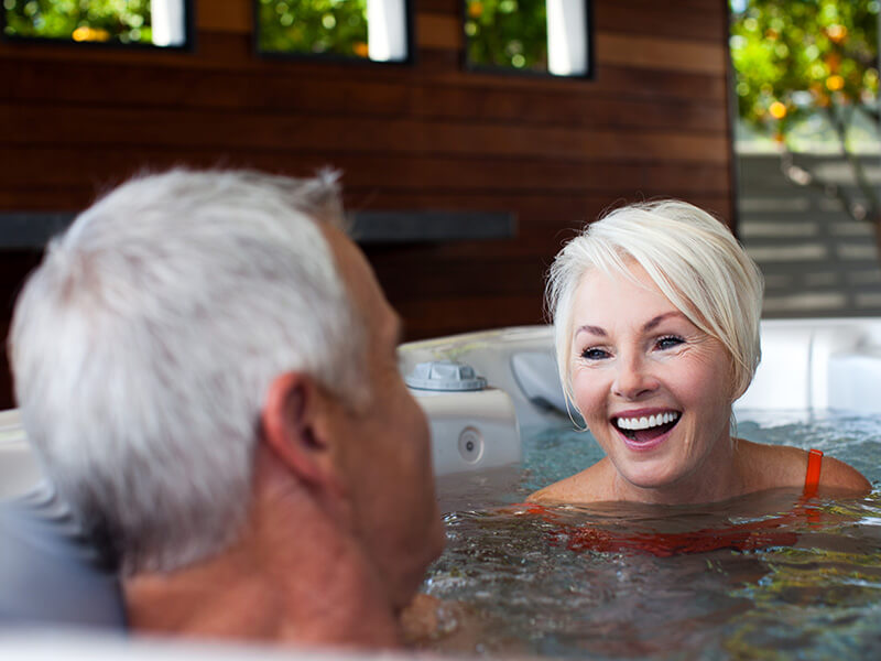 Smiling older couple enjoying a conversation while relaxing in a warm hot tub, with a modern wooden-paneled outdoor space in the background.