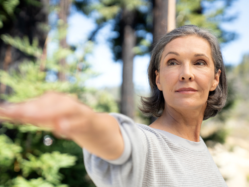 Woman practicing a yoga pose outdoors, extending her arm with focus and calm expression, surrounded by trees and natural light.