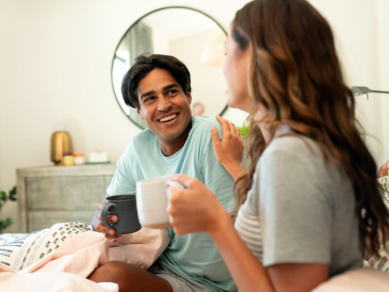 Smiling man holding a mug and talking with a woman in a cozy bedroom setting, suggesting a relaxed morning conversation